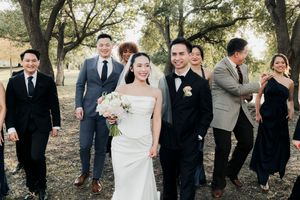 Joyful bride and groom hold hands while walking with wedding party through sunlit outdoor setting.