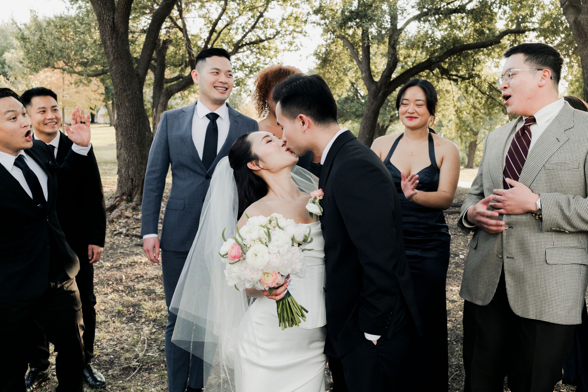 Bride and groom share romantic kiss surrounded by cheering wedding party under oak trees.