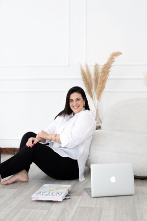 Relaxed person in white shirt and black pants sits on floor with laptop and pampas grass decor in minimalist white room.