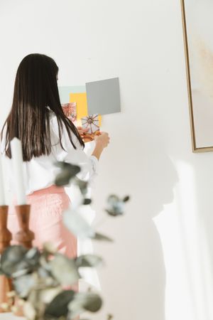 A person in a white top and dark hair holds up paint color swatches against a white wall with eucalyptus branches visible.