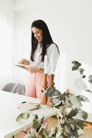 Woman in white sweater works at desk with pink chair and eucalyptus plant in bright modern workspace.