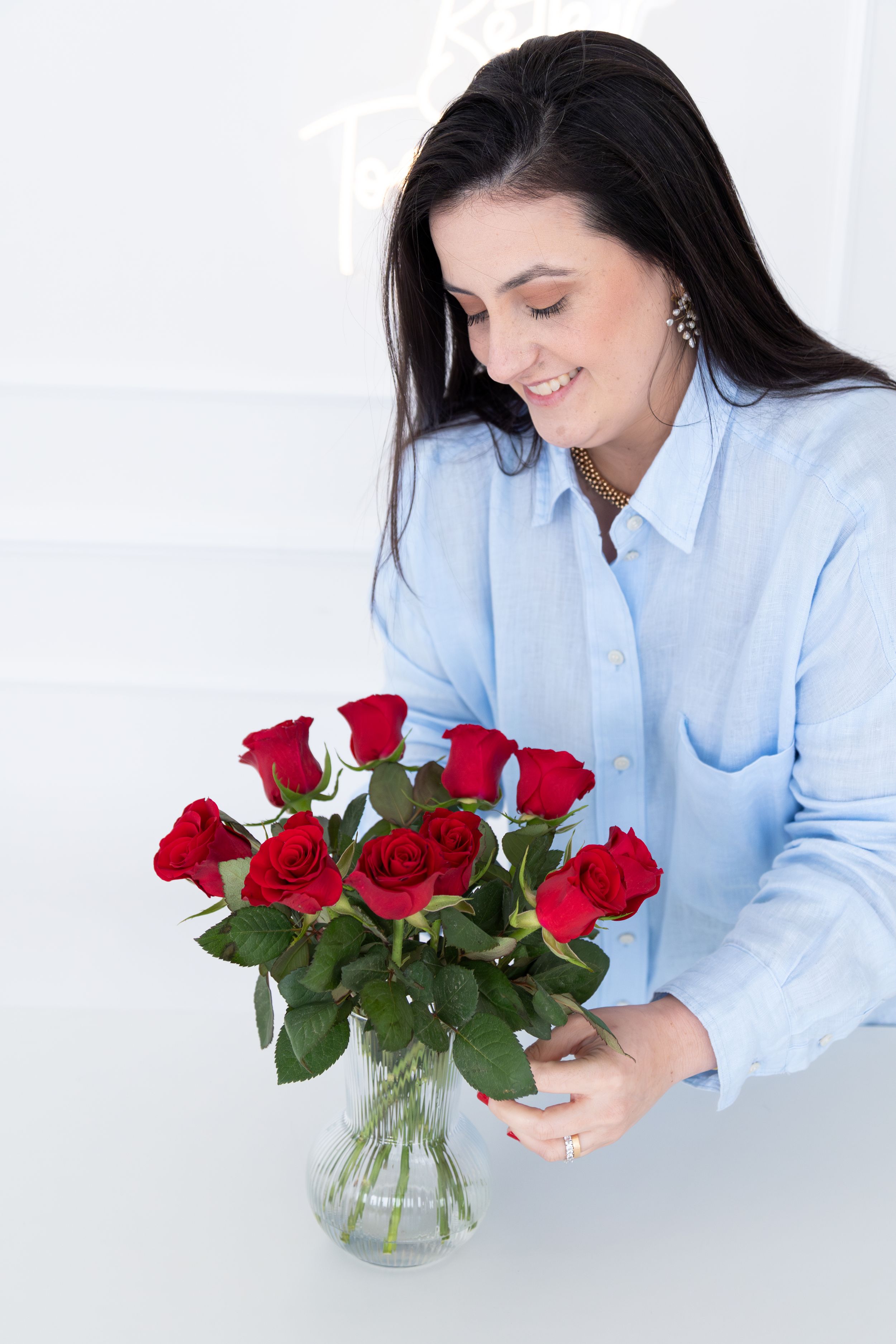 Woman in light blue shirt arranges fresh red roses in clear glass vase against white background.