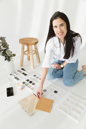 Designer in denim overalls sits on floor reviewing printed layouts and wooden furniture pieces in bright studio setting.