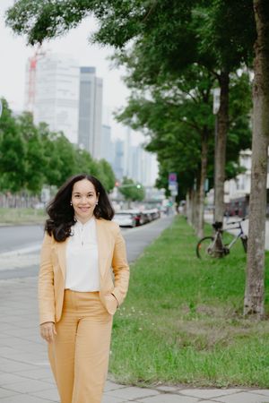 A person in a yellow pantsuit and white top poses on a tree-lined urban sidewalk with city buildings in the background.