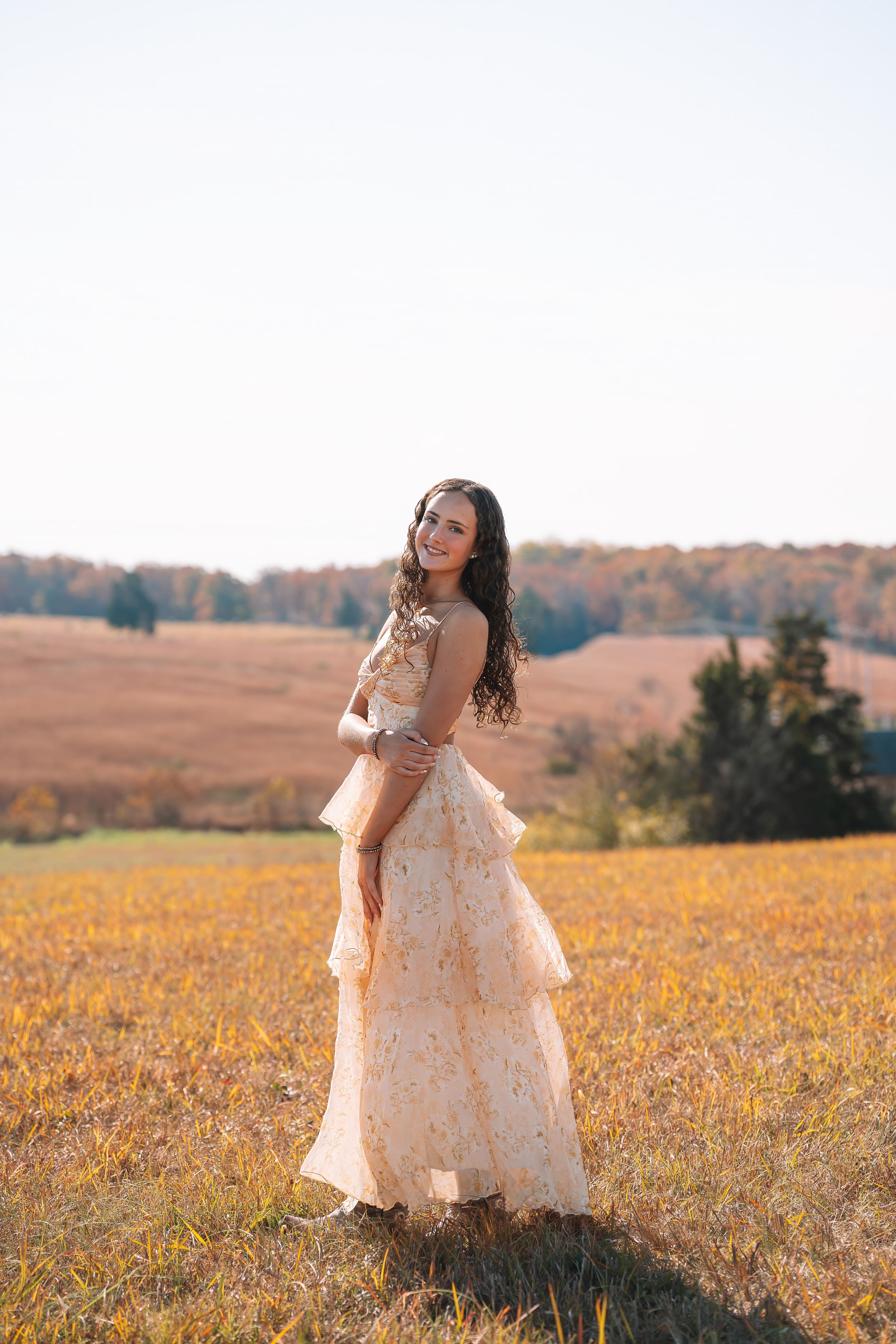 A young woman wearing a long, lace dress stands in a golden field, with a forest in the background.