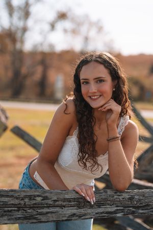 A young woman with long curly hair smiling and leaning on a wooden fence in a rural setting.