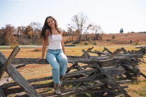 A young woman stands on a wooden fence in a rural field on an autumn day.