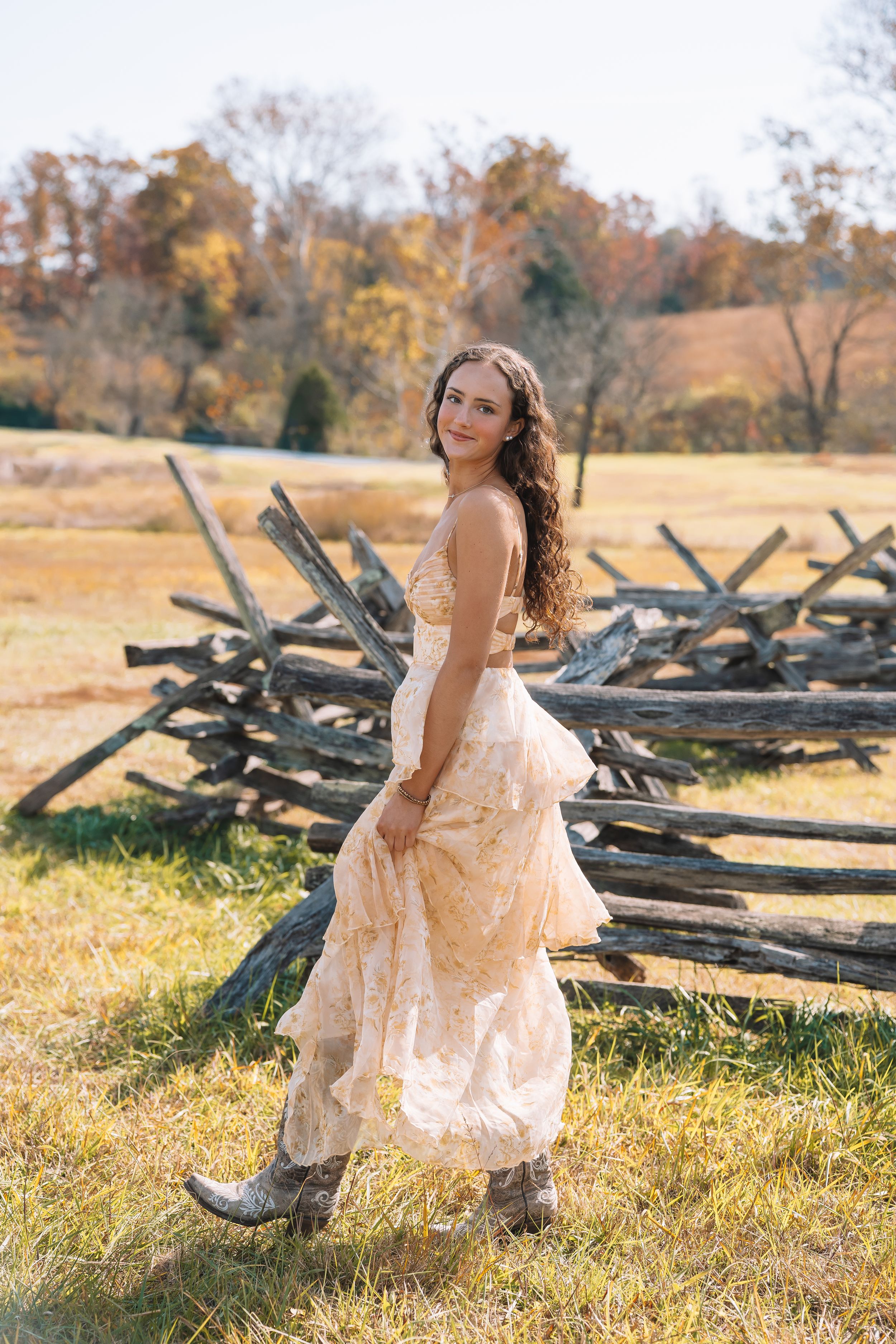 A woman in a long, flowing dress stands in a field with a wooden fence in the background.