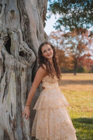 A young woman with long curly hair stands next to a large tree trunk in a grassy field on an autumn day.