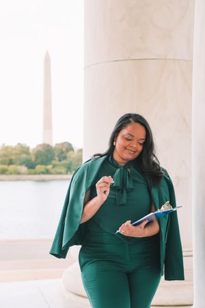 A woman in a green outfit stands in front of a white column, smiling and holding a tablet.