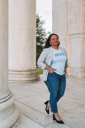 A smiling woman wearing a blue shirt and jeans stands in front of stone columns.