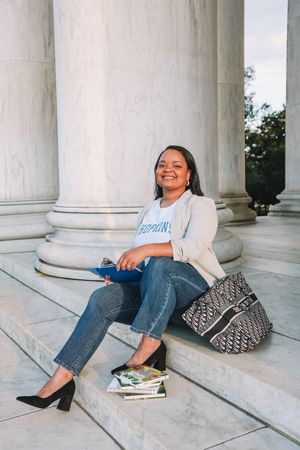 A smiling woman wearing a white shirt and jeans sits on the steps of a building with columns.