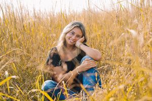 A young woman sits in a field of tall grass, smiling and petting a small dog beside her.