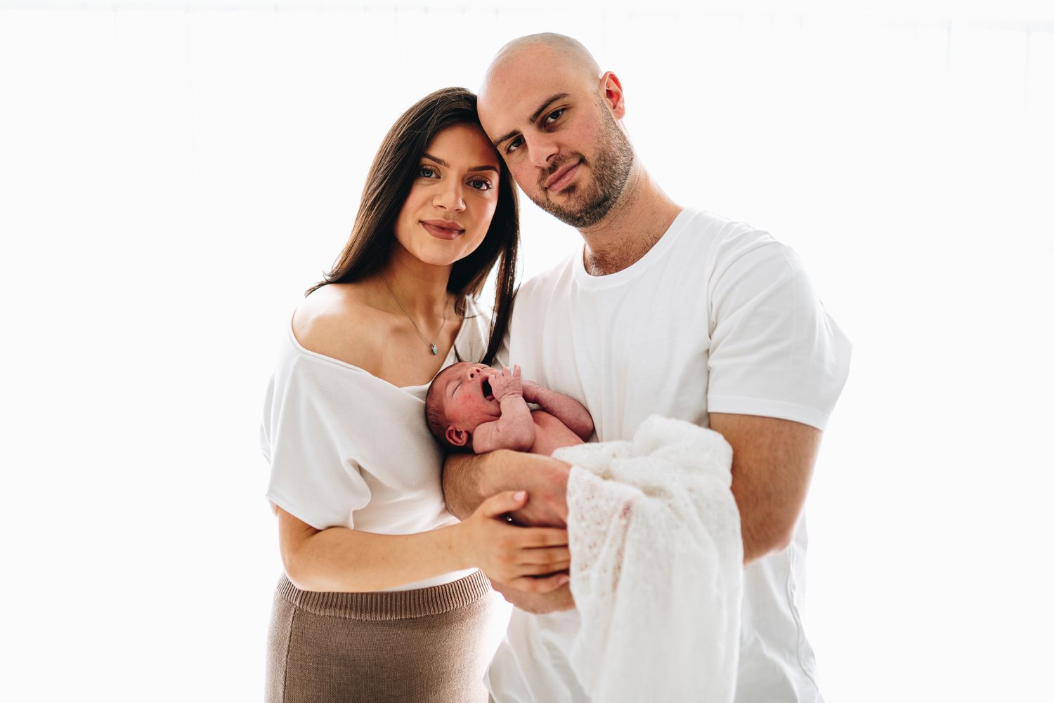 A young couple in white tops smiles while holding their newborn baby against a white background taken by Melbourne newborn photographer.