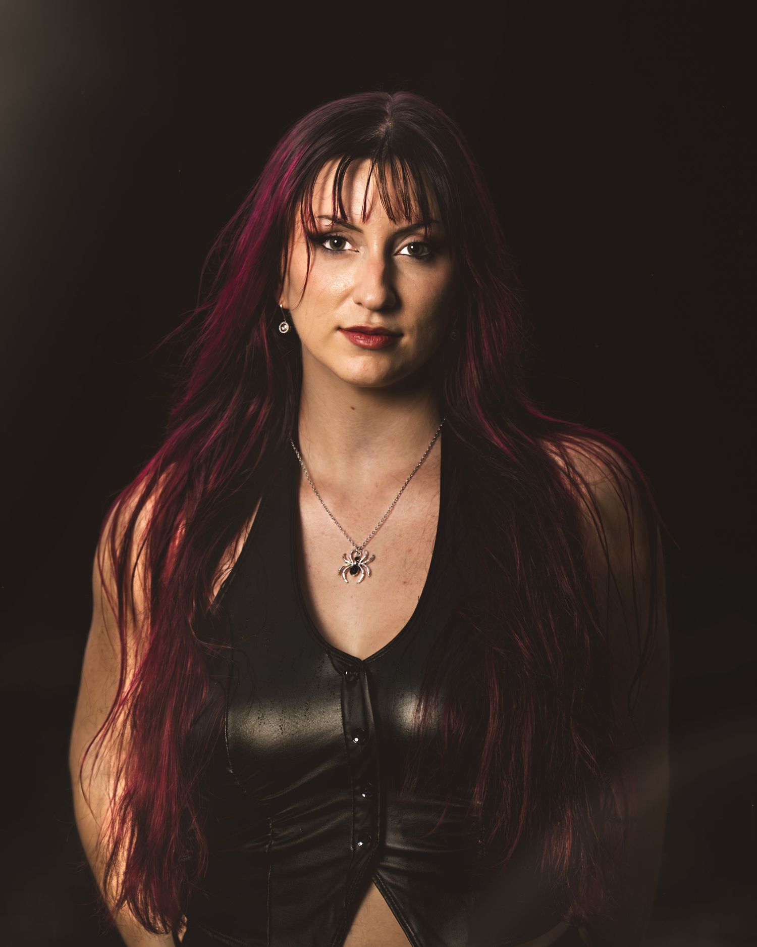 Woman with long red hair wearing black leather top and spider necklace poses against dark background.