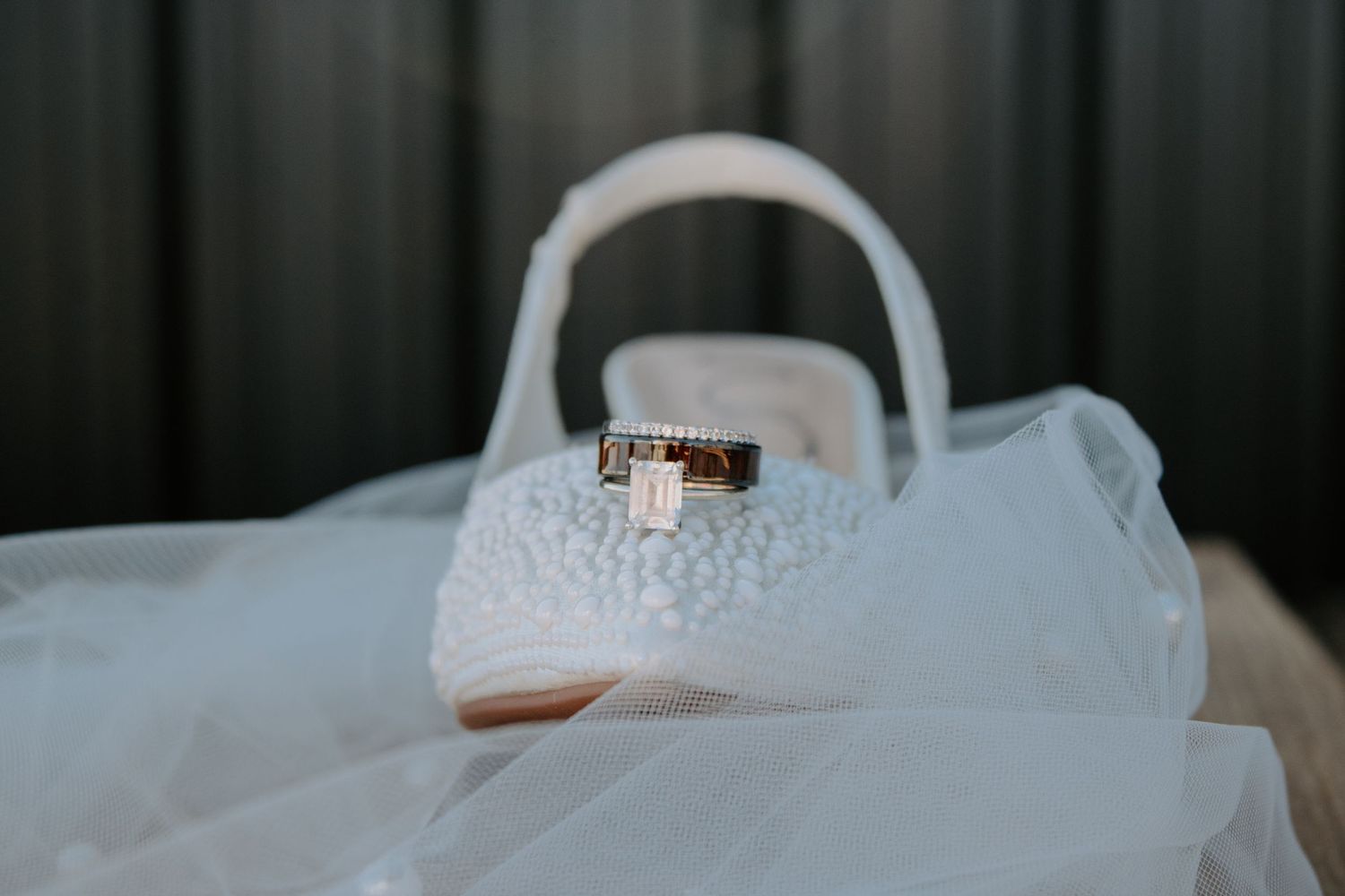 Wedding rings resting on a white lace bridal shoe with veil and flower girl basket in the background.