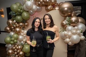 Two women in black outfits holding cocktails and smiling in front of an elegant balloon arch decoration.