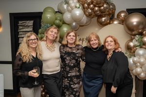 Five women smile together under a balloon arch at a festive indoor celebration event.