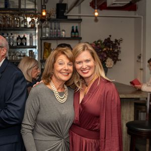 Two smiling women pose together at a bar, one wearing pearls and gray, the other in burgundy.