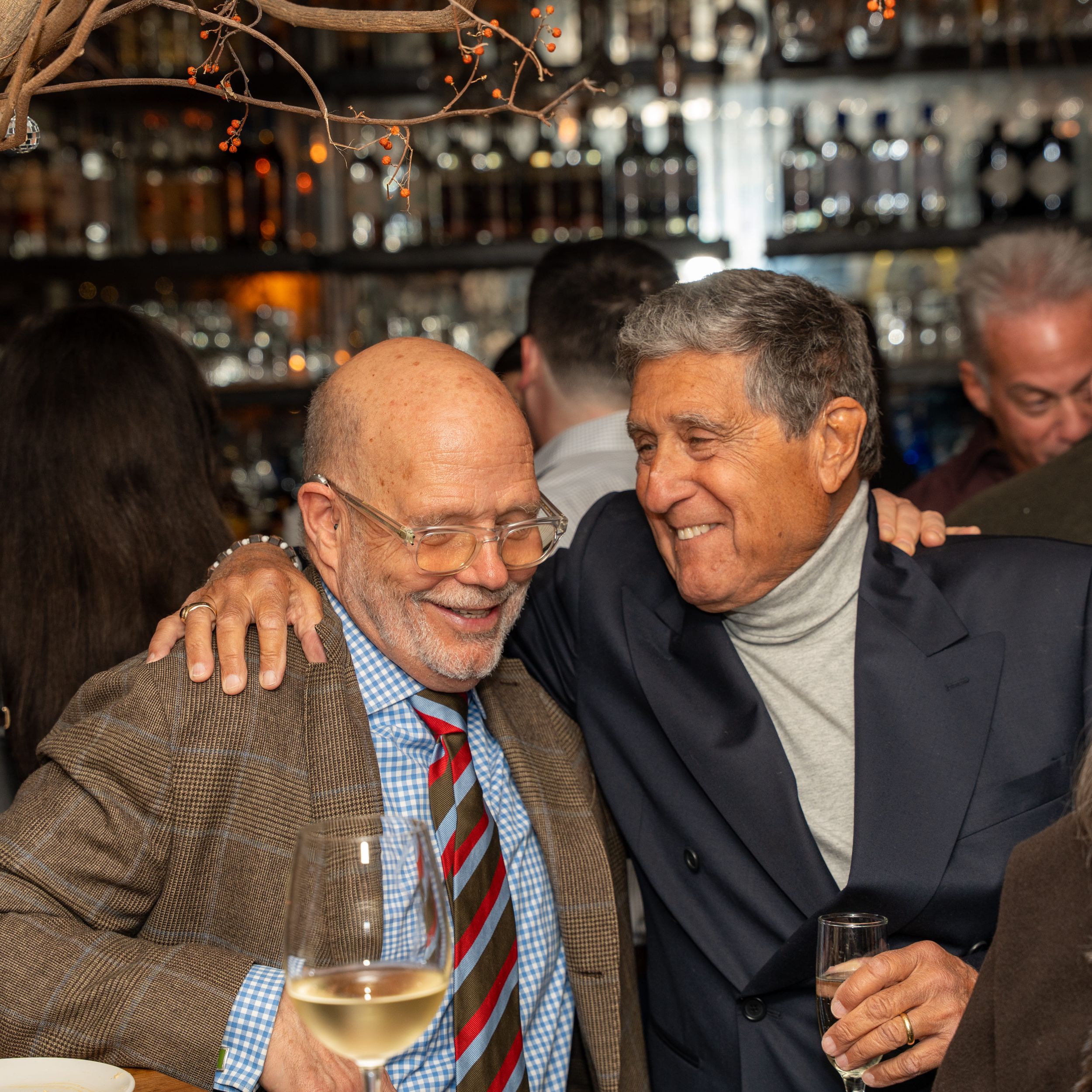 Two older men sharing a warm embrace and laughing together at an upscale bar event with drinks in hand.