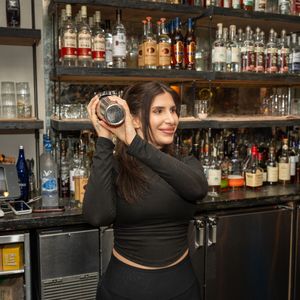 Female bartender posing with a cocktail shaker at a well-stocked bar.