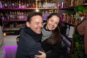 A man and woman laugh and embrace playfully at a bar with bottle-lined shelves behind them.