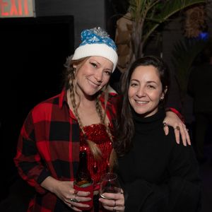 Two women smile at a festive holiday party, one wearing a plaid shirt and winter hat.