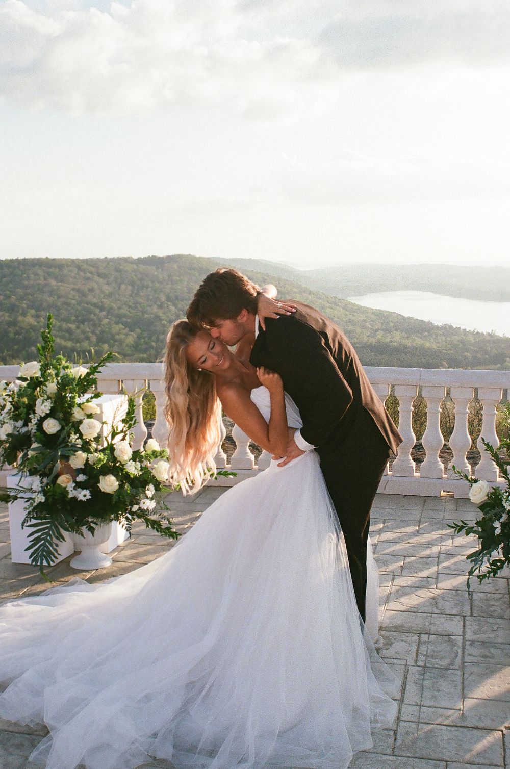 Groom embraces bride in strapless tulle gown beside white floral arrangements on terrace with lake view.