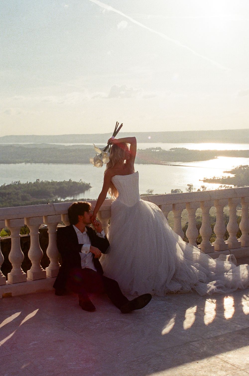 Bride in flowing tulle gown poses dramatically at sunset on balcony overlooking shimmering lake landscape.