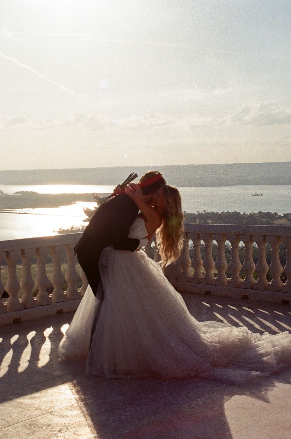 Couple shares romantic dip kiss on sun-drenched terrace balcony with breathtaking river and sky backdrop.