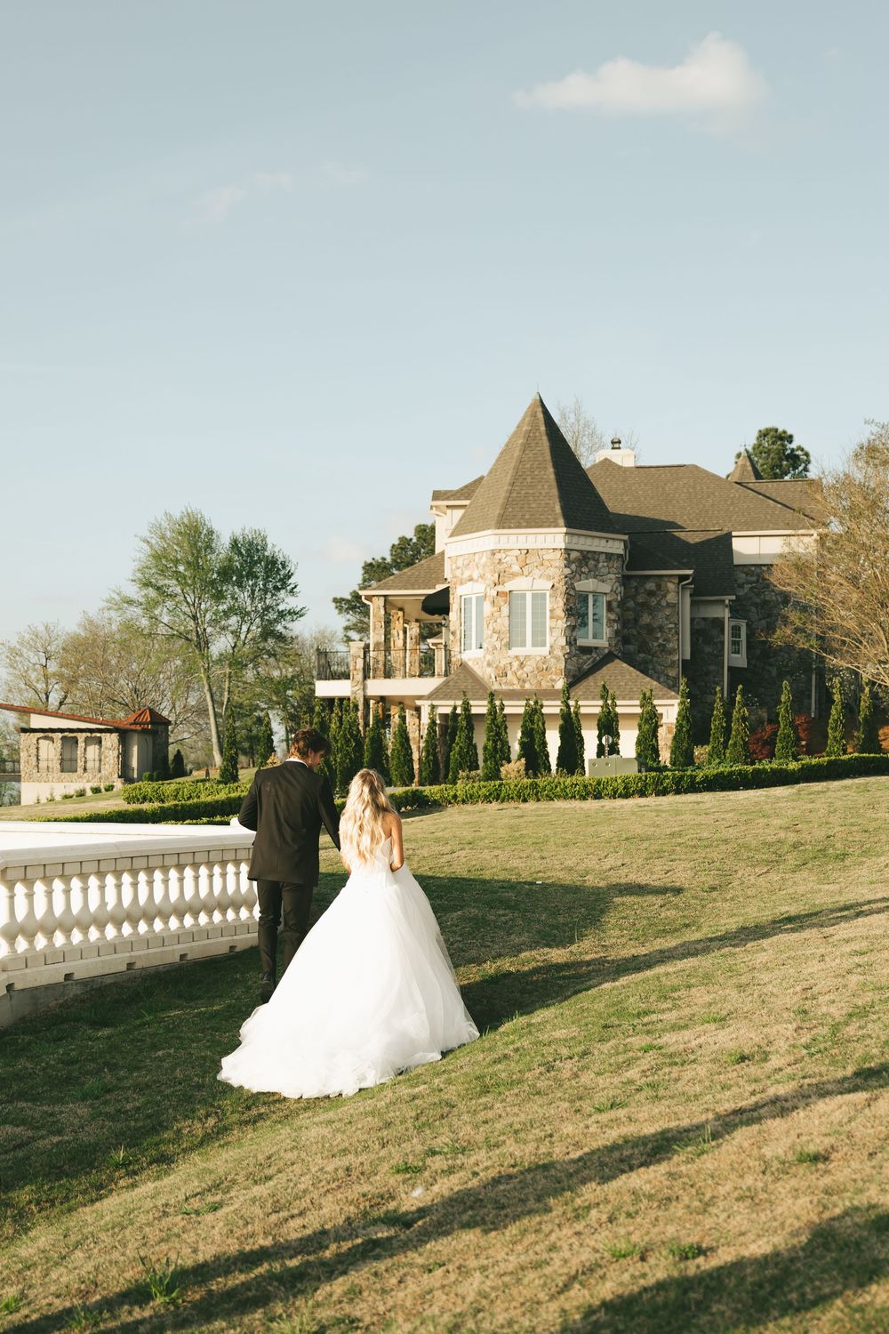 Bride and groom walking toward elegant stone mansion on sunny wedding day.