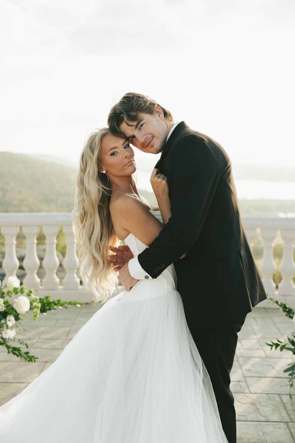 Elegant couple poses on stone balcony, bride in layered tulle gown embracing groom in black tuxedo.