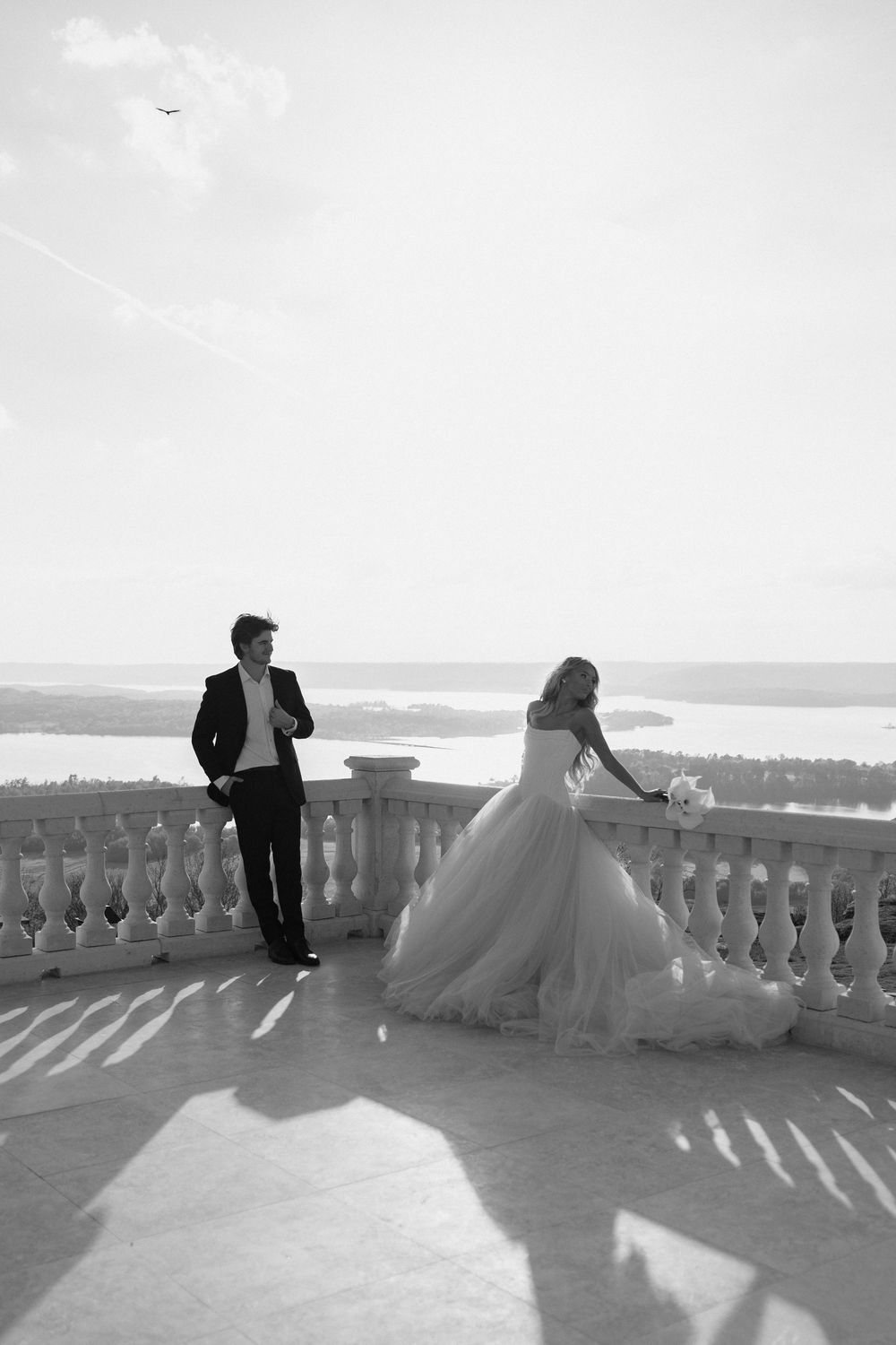 Black and white photo of bride and groom standing apart on grand balcony overlooking scenic water.
