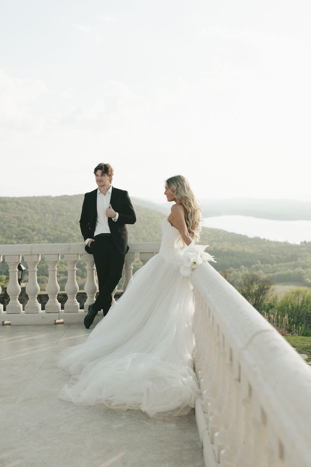Bride holding white bouquet in strapless tulle gown stands with groom on balcony with mountain views.
