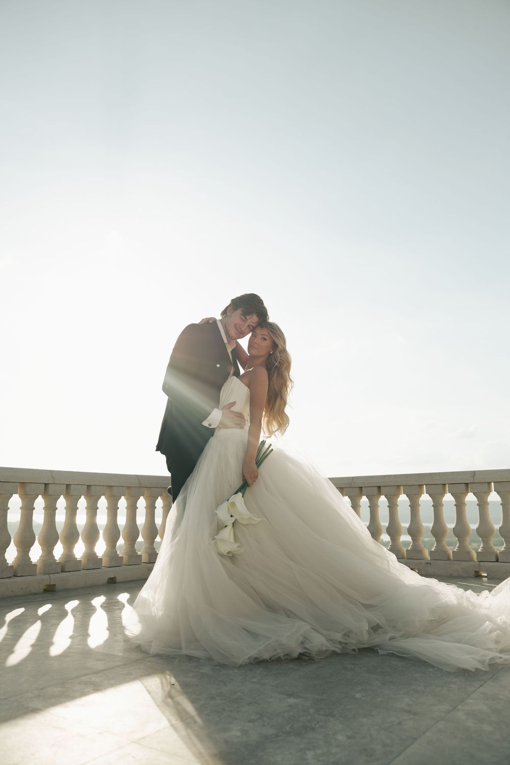 Bride and groom share romantic kiss on sunlit stone balcony, bride in flowing white tulle gown.