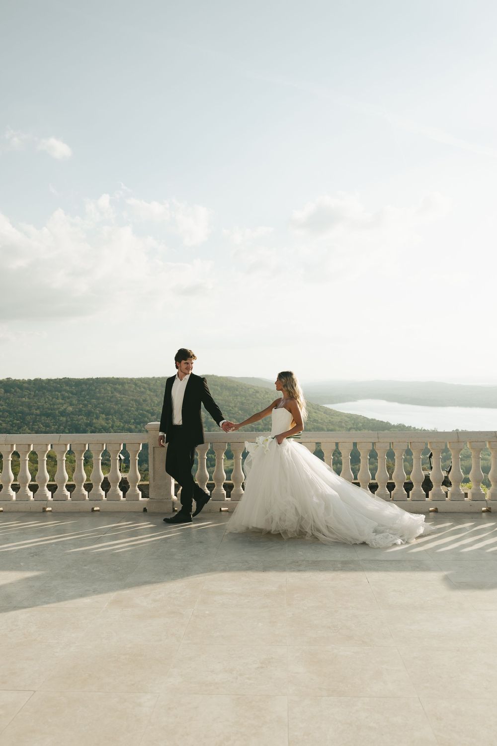 Bride and groom hold hands on elegant stone terrace overlooking lush green hills and serene lake.