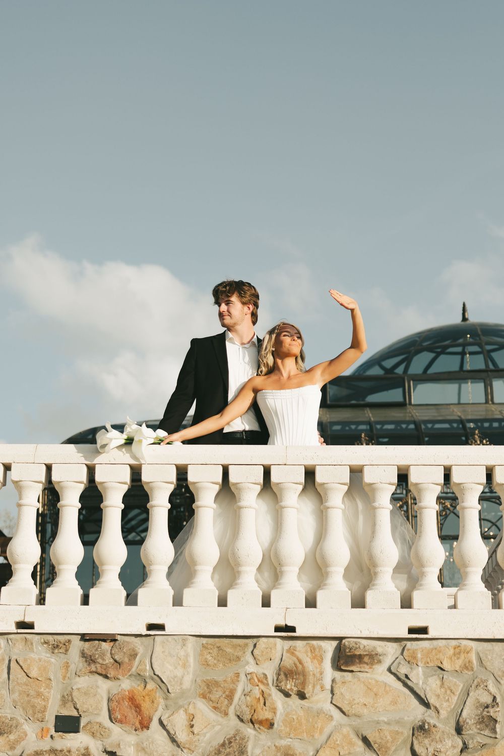 Bride in strapless white gown waves joyfully beside groom on white balustrade under blue sky.