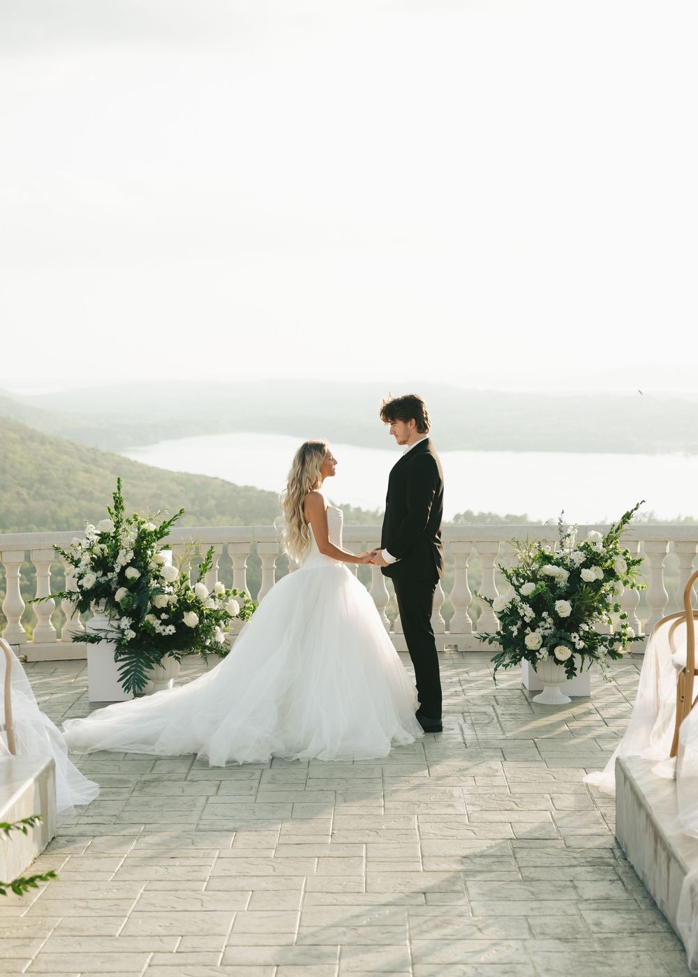 Bride and groom exchange vows on scenic terrace surrounded by white floral arrangements and lake views.
