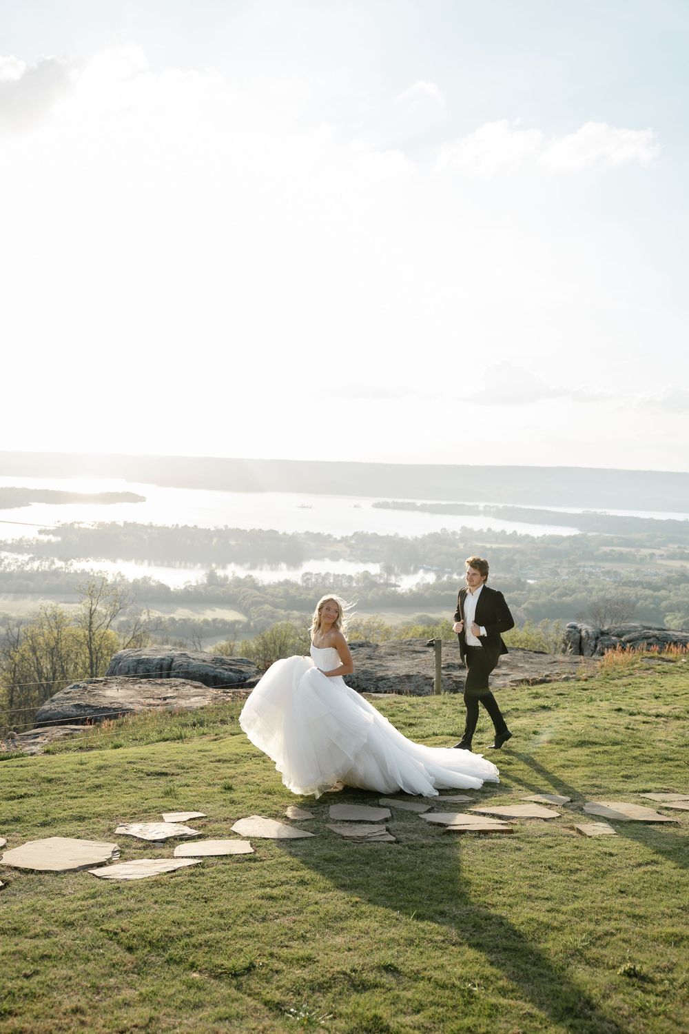 Bride in flowing white ballgown with long train walks with groom in black tuxedo on scenic hilltop overlooking water.