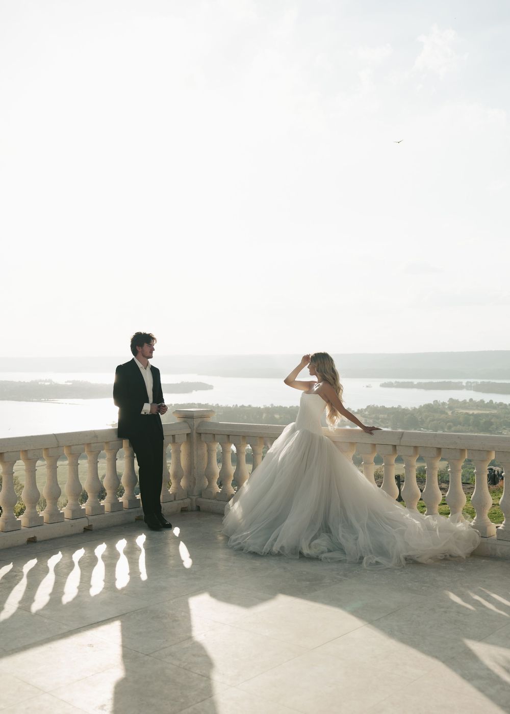 Bride in dramatic tulle gown poses on sunlit terrace balcony with panoramic lake and sky views.