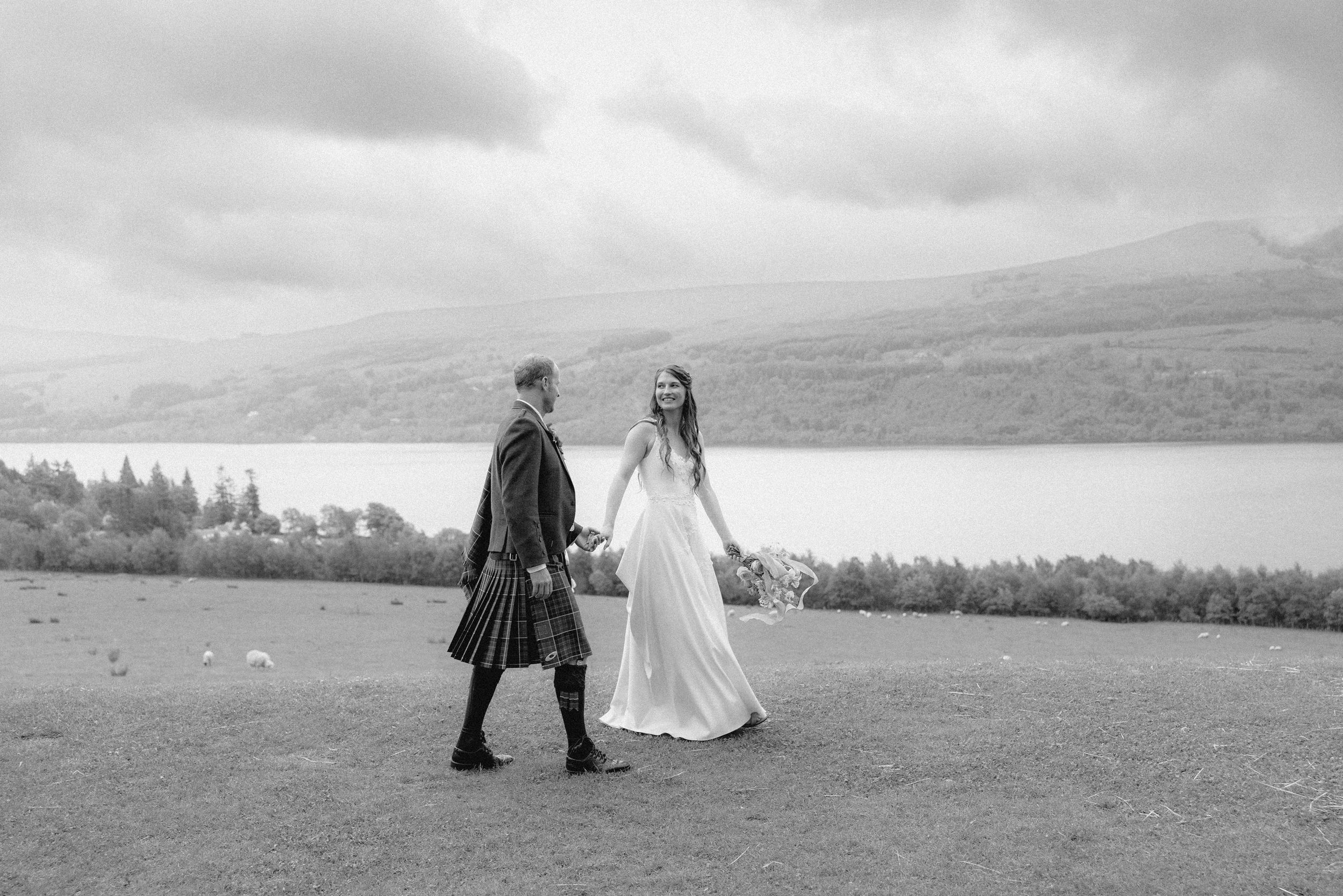 A couple in wedding attire walks along a scenic mountain landscape in black and white photography.