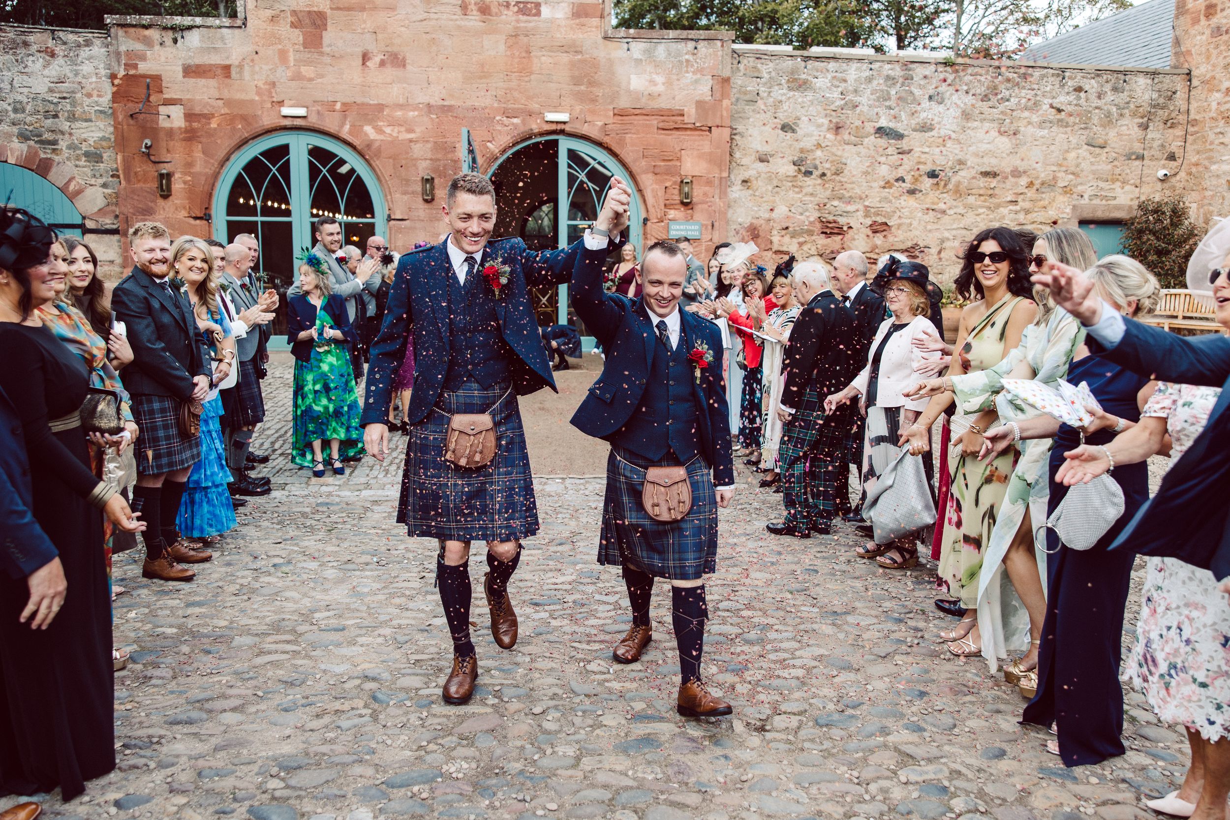 Wedding guests celebrate by throwing confetti on a cobblestone courtyard outside a historic stone building.