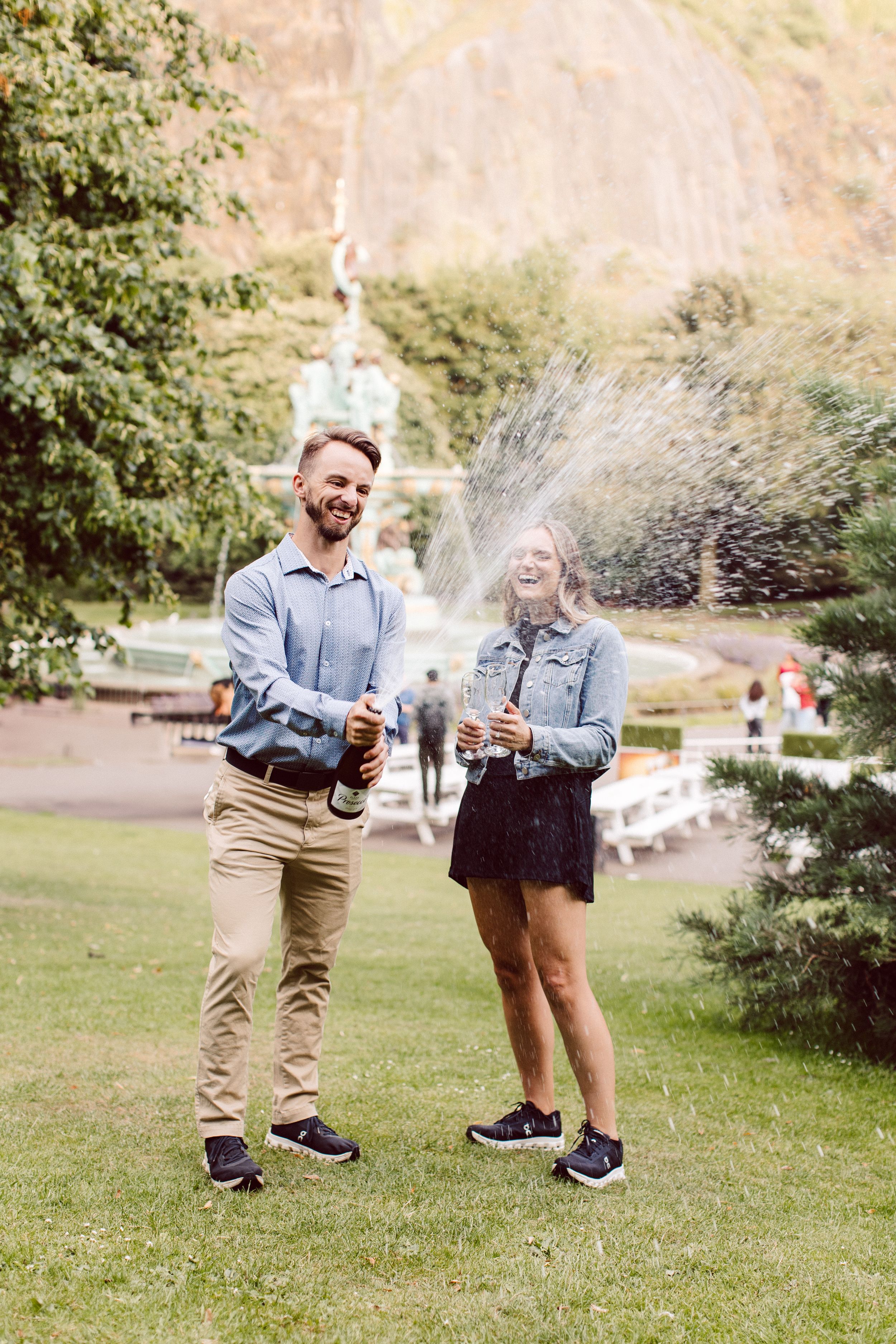 Two people laugh and spray champagne bottles outdoors on a grassy lawn with trees and mountains in the background.