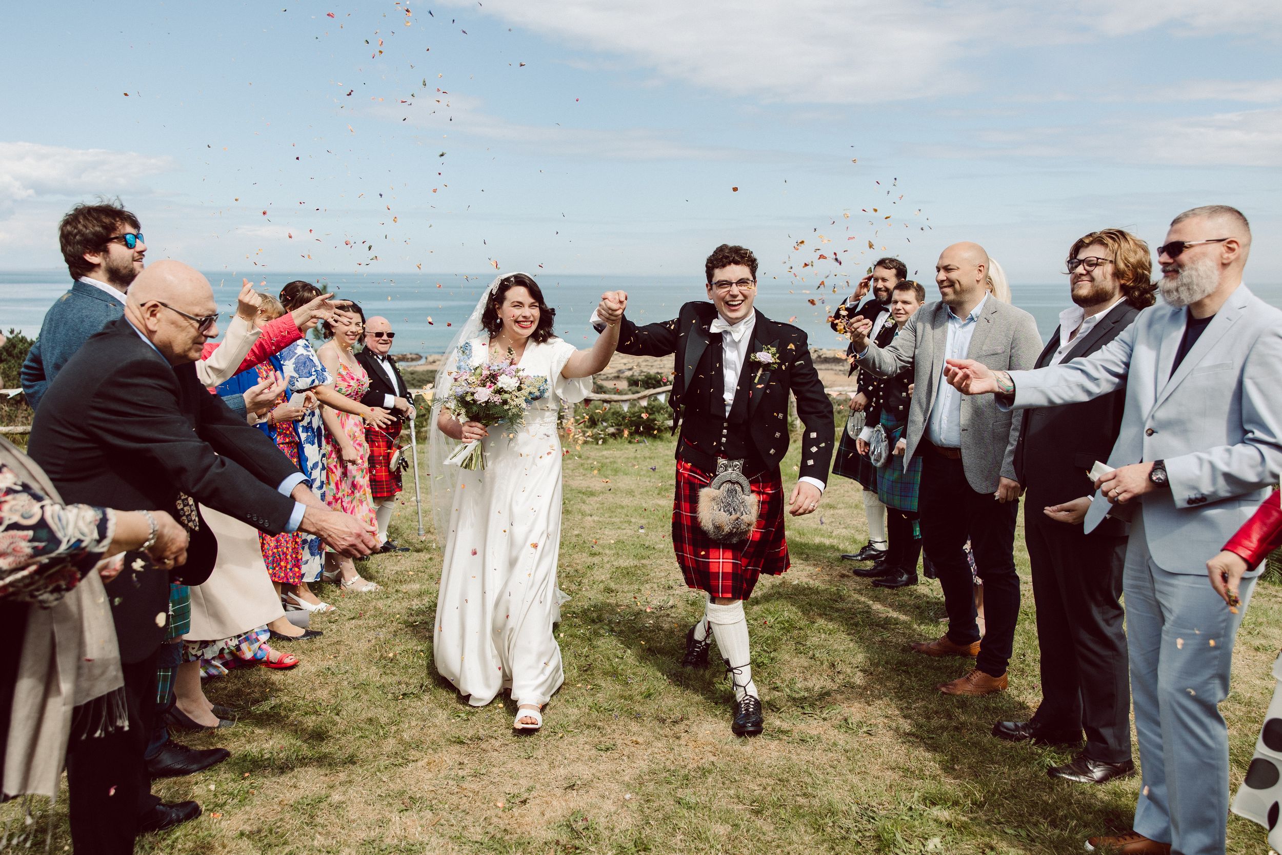 Wedding guests form a joyful receiving line outdoors on a grassy field to celebrate the newlyweds.