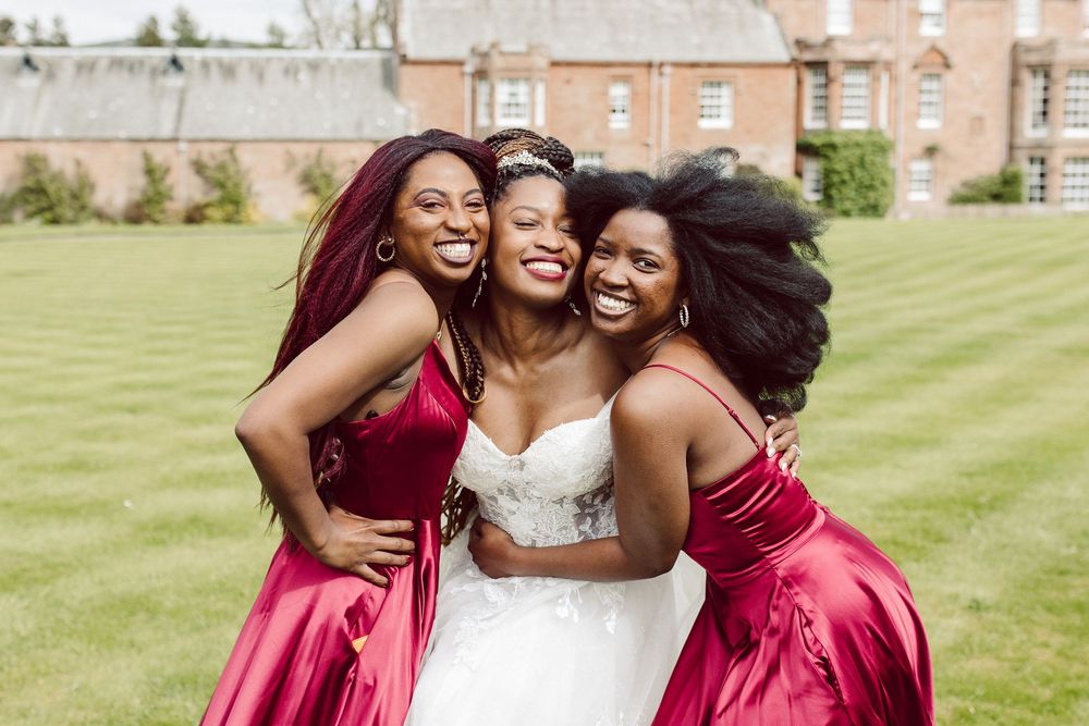 Bridesmaids in burgundy dresses pose with bride in white gown on green lawn during outdoor wedding celebration.