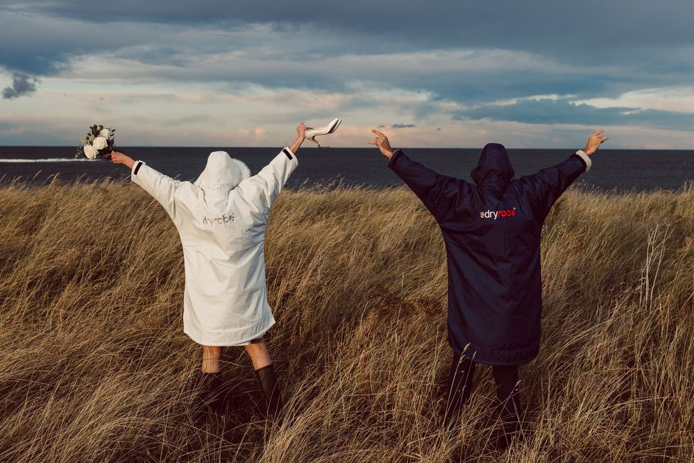 Two people in hooded robes stand with raised arms in tall grass against a dramatic cloudy sky at sunset.