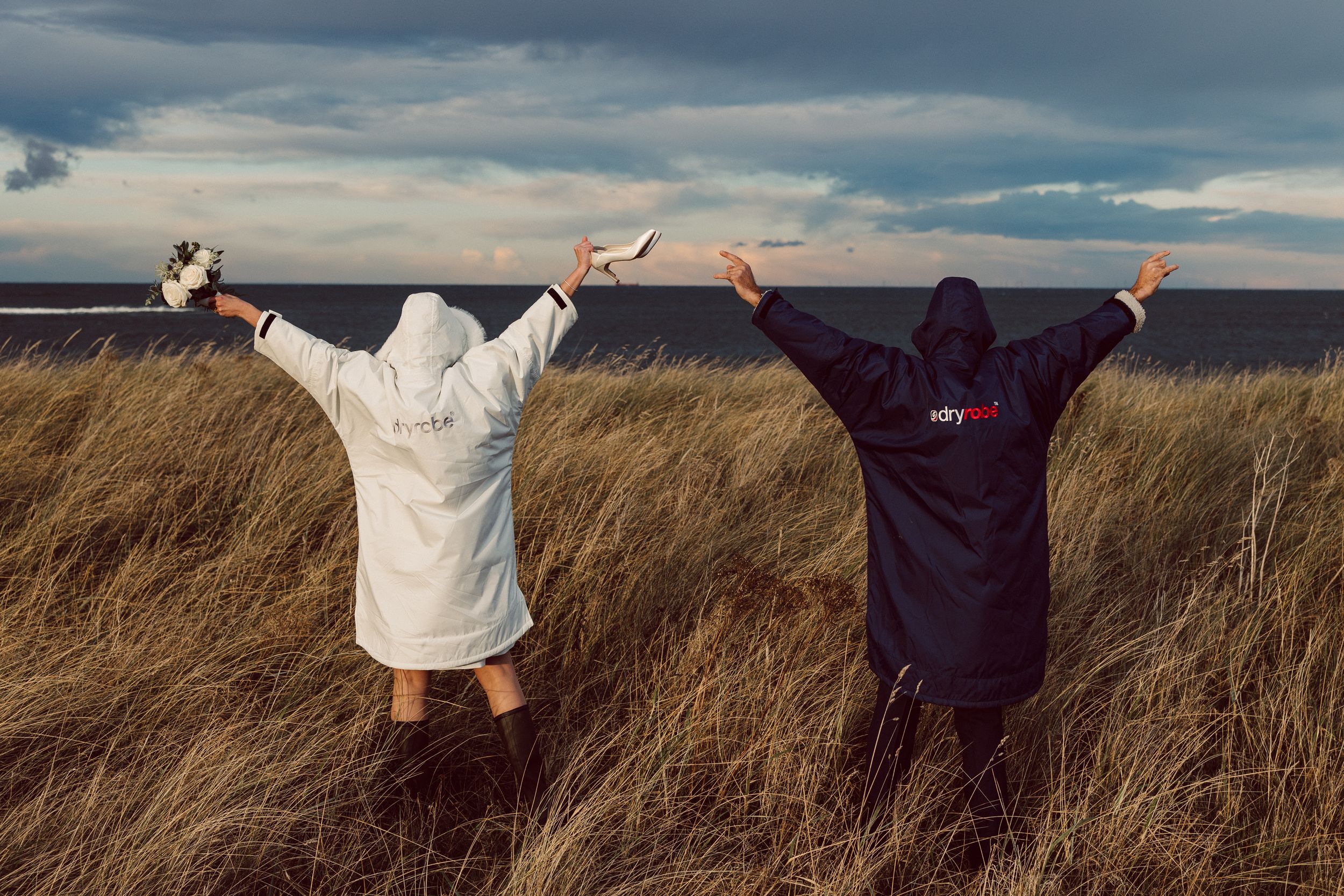 Two people in hooded robes stand with raised arms in tall grass against a dramatic cloudy sky at sunset.