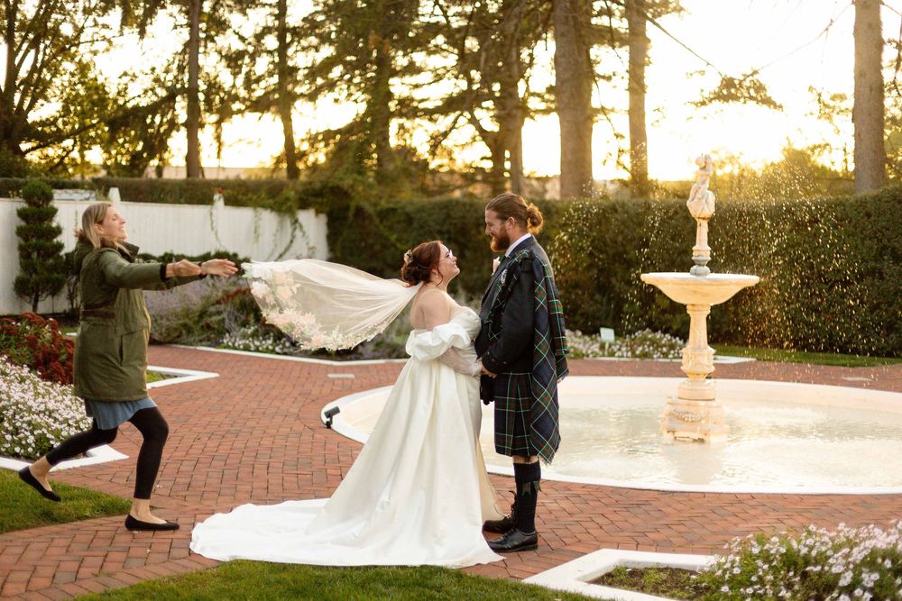 Wedding couple stands near fountain at sunset while photographer captures their special moment in garden setting.