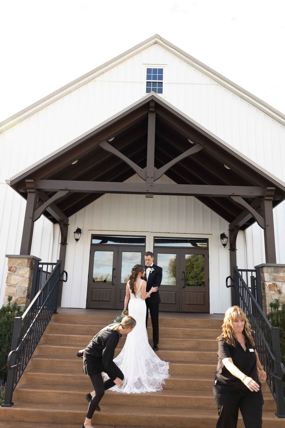 Wedding party stands on steps of a white barn venue with rustic wooden beams and stone pillars.