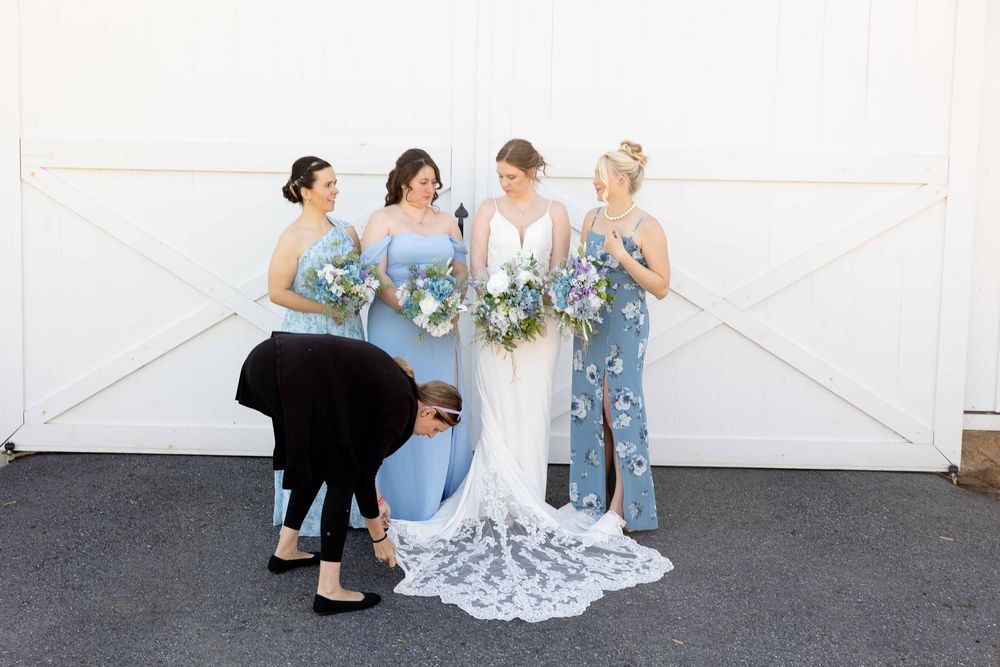 A group in blue dresses and gowns gather for a wedding photo against a white door while someone adjusts the dress train.
